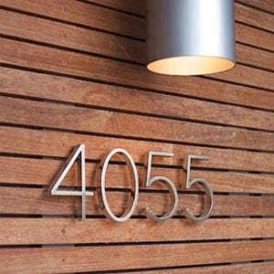 May include: Silver metal house numbers, "4055", are mounted on a horizontal wooden plank wall. A cylindrical silver light fixture is mounted above the numbers. The wood has a warm, natural tone.