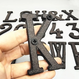 May include: A dark brown, cast iron letter "K" is held in a hand, with other letters and numbers in the background. The letter is textured and has a screw in the center. The other letters and numbers are also dark brown.