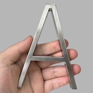 May include: A brushed metal letter "A" is held in a hand against a gray background. The capital letter is a solid, three-dimensional design with a modern, minimalist aesthetic. The metal has a textured, matte finish.