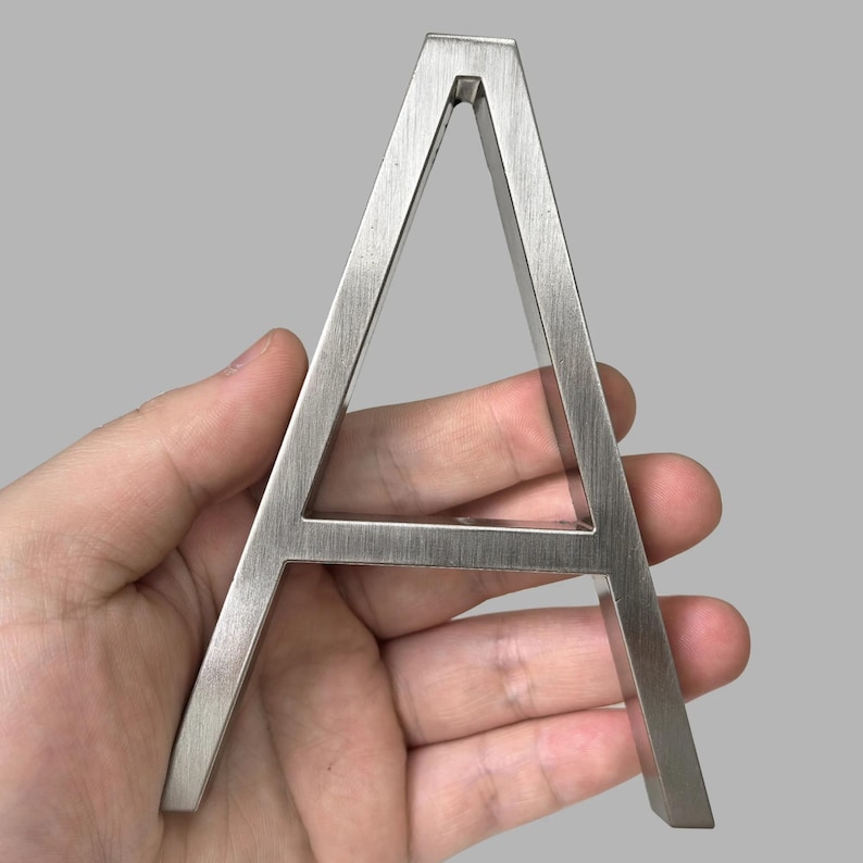 May include: A brushed metal letter "A" is held in a hand against a gray background. The capital letter is a solid, three-dimensional design with a modern, minimalist aesthetic. The metal has a textured, matte finish.