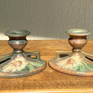 May include: Two ceramic candle holders with a floral design. The holders are brown with green and yellow accents. Each has a circular base with a flower and leaf pattern, and a cylindrical top for holding a candle.