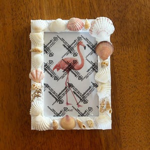 May include: White rectangular photo frame decorated with various seashells. The frame holds a picture of a pink flamingo. The frame is designed for a 10x15 cm photo.
