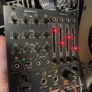May include: A black modular synthesiser module featuring a parametric EQ section with multiple knobs, an output mixer with four vertical faders illuminated by red LEDs, and various input and output jacks labelled with white text and numerical markings.