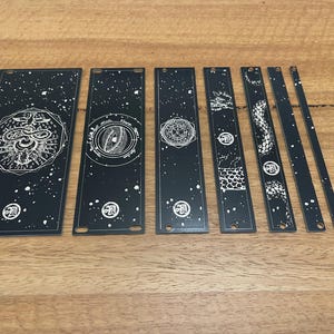May include: A set of black metal panels featuring white celestial and geometric designs. The panels are of various sizes and shapes, with a starry background and a circular logo. The designs include a snake, eye, and other symbols.