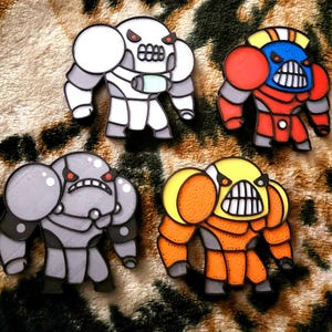 May include: Four enamel pins featuring stylized, muscular armored figures with wide, toothy grins and red eyes. The figures are colored in white, red and blue, solid grey, and orange and yellow, each outlined in thick black lines.