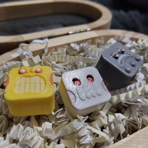 May include: Three colorful, square-shaped decorative items with cartoon-like faces. One is yellow with a toothy grin, another is white with a skull design, and the third is gray with red accents. They rest in a wooden box filled with paper shreds.