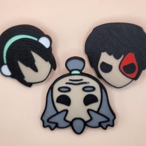 Könnte beinhalten: Drei dekorative Gegenstände in Form von Charakteren. Einer hat schwarzes Haar mit einem grünen Stirnband und weißen Akzenten. Ein anderer hat schwarzes Haar und ein rotes Mal im Gesicht. Der dritte hat graues Haar und ein blaues Kopfstück. Alle haben hellbraune Gesichter.