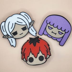 Puede incluir: Tres pines de personajes de dibujos animados. Uno tiene el pelo blanco y orejas puntiagudas, otro tiene el pelo morado y el tercero tiene el pelo rojo. Todos tienen contornos negros y expresiones neutras. El fondo es de color melocotón claro.