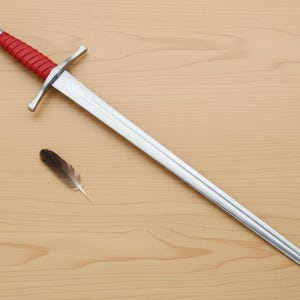 May include: A medieval-style sword with a silver blade and crossguard, and a red wrapped handle. A small feather rests near the sword on a light wood surface. The sword is a decorative item.
