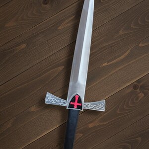 May include: A silver-colored sword with a pointed blade and a black handle. The crossguard features a red cross on a shield. The hilt has a red gem at the end. The sword is set against a dark wood background.