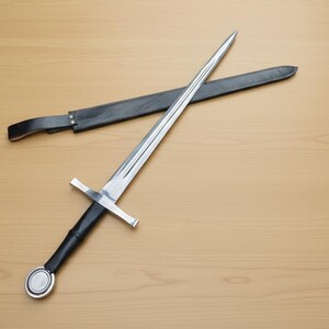May include: A long, silver sword with a cross-shaped hilt and a black handle, resting on a wooden surface. A black leather sheath lies nearby. The sword's blade is sharp and reflective.