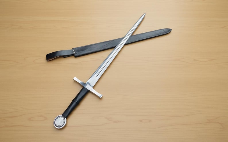 May include: A long, silver sword with a cross-shaped hilt and a black handle, resting on a wooden surface. A black leather sheath lies nearby. The sword's blade is sharp and reflective.