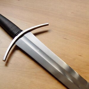 May include: A medieval-style sword with a silver blade and a black wrapped handle. The sword features a crossguard and a rounded pommel. The blade is angled towards the bottom right, set against a wooden background.