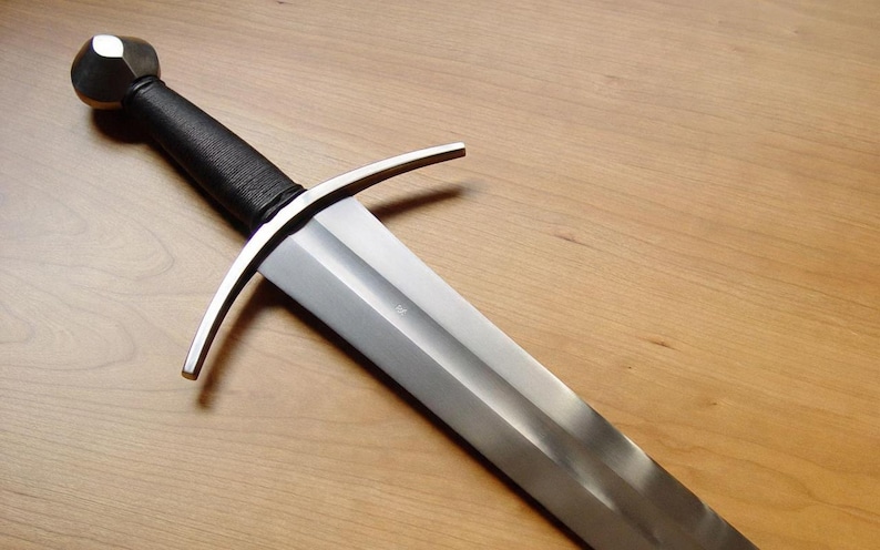 May include: A medieval-style sword with a silver blade and a black wrapped handle. The sword features a crossguard and a rounded pommel. The blade is angled towards the bottom right, set against a wooden background.