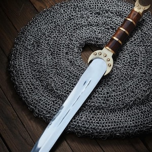 May include: A Viking-style dagger with a polished steel blade and a wooden handle accented with brass. The dagger rests on a circular piece of chainmail, set against a dark wooden background. The handle features a crescent-shaped brass guard.