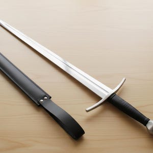 May include: A long, silver sword with a black handle and a silver pommel, next to a black leather sheath. The sword has a crossguard and a pointed tip. The sheath is rectangular with a strap.