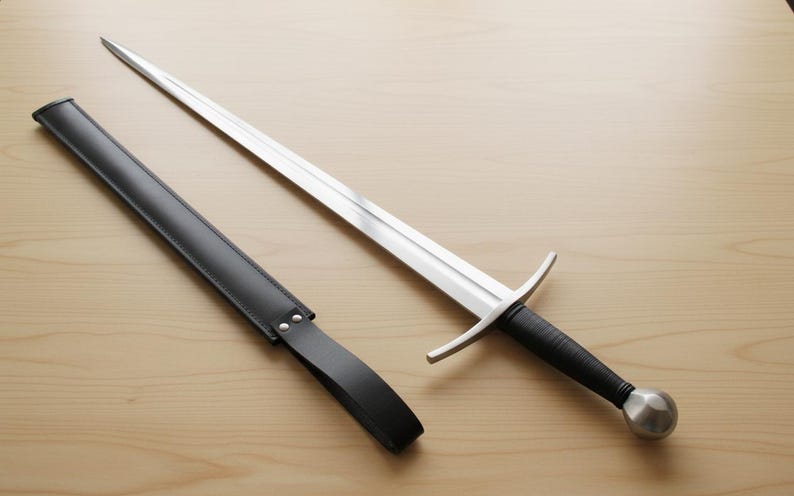 May include: A long, silver sword with a black handle and a silver pommel, next to a black leather sheath. The sword has a crossguard and a pointed tip. The sheath is rectangular with a strap.