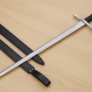 May include: A medieval-style sword with a silver blade and a black handle. The sword is laid across a black leather sheath on a light wood surface. The sword's crossguard and pommel are silver.