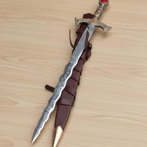 May include: A decorative sword with a silver blade and ornate gold hilt, resting in a brown leather sheath. The blade has etched designs and text. The hilt features a red gem. The sheath has a gold-colored tip.
