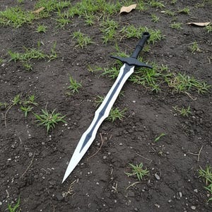 May include: A black and white sword with a long, pointed blade. The sword has a black handle and crossguard. The blade is white with a black design down the center. The sword is lying on the ground.