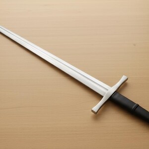 May include: A long, silver sword with a cross-shaped hilt and a black handle. The sword is lying on a light brown wooden surface. The blade is sharp and pointed, and the handle has a rounded pommel.