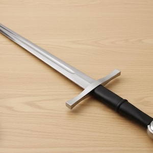 May include: A medieval sword with a silver blade and a black handle, on a wooden surface. The sword is accompanied by a black leather sheath. The sword's length is approximately 90 cm.