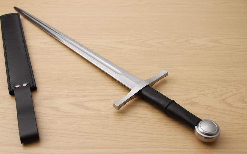 May include: A medieval-style sword with a silver blade and a black handle, resting on a wooden surface. A black leather sheath is positioned to the left of the sword.