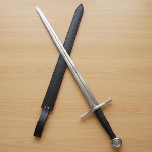 May include: A medieval sword with a silver blade and a black handle, resting on a wooden surface. The sword is crossed with a black leather sheath. The design features a crossguard and a rounded pommel.