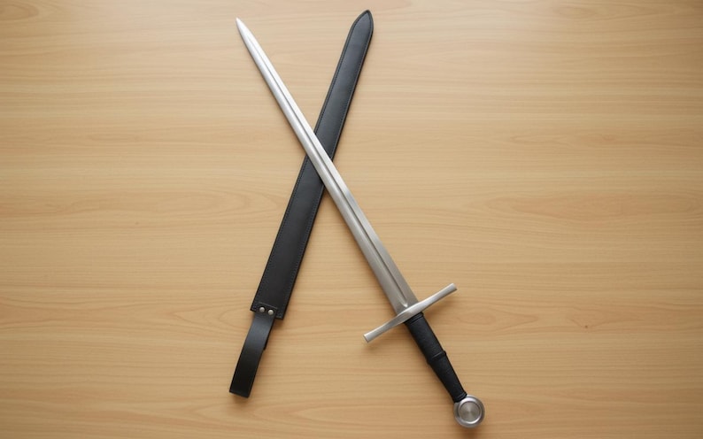 May include: A medieval sword with a silver blade and a black handle, resting on a wooden surface. The sword is crossed with a black leather sheath. The design features a crossguard and a rounded pommel.