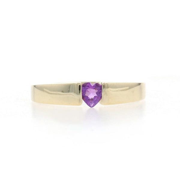 Puede incluir: Un anillo de banda dorada que presenta una única piedra preciosa morada en forma de corazón engastada en el centro. La banda tiene un acabado liso y pulido con un perfil plano, y la piedra está sujeta entre dos secciones de la banda de oro.