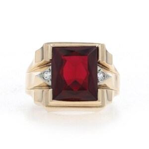 Peut inclure: Une bague en or ornée d'une grande pierre précieuse rectangulaire rouge foncé au centre. L'anneau présente un design géométrique structuré avec une petite pierre claire de taille ronde incrustée de chaque côté du sertissage central.