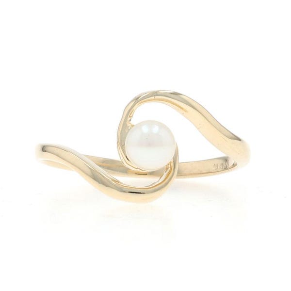 Puede incluir: Un anillo de oro con una perla blanca redonda centrada entre dos bandas curvas de oro amarillo pulido que se encuentran para sostener la perla en un diseño de estilo bypass, creando una silueta metálica suave y continua.