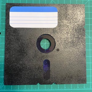 May include: A black 3.5-inch floppy disk with a textured surface. It features a label area with blue and white stripes, a central circular opening, and a rectangular slot. The disk is placed on a green grid.
