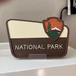 May include: Wooden National Park sign with a cream-colored top section and a brown bottom section. The sign features the National Park Service logo in the upper right corner, depicting a mountain, tree, and sunset. The words "NATIONAL PARK" are printed in white.