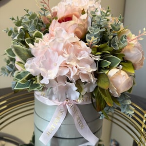 May include: A floral arrangement featuring light pink peonies and hydrangeas, accented with green foliage. The flowers are arranged in a round, light green box adorned with a pink ribbon that reads "JUST FOR YOU". The arrangement sits on a reflective surface.