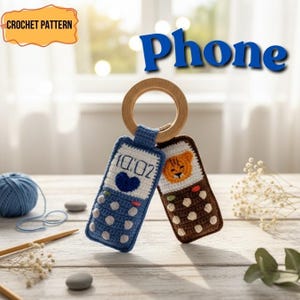 Puede incluir: Patrón de ganchillo para dos teléfonos de juguete, uno azul y otro marrón, unidos a un anillo de madera. El teléfono azul tiene una pantalla blanca con un corazón y la hora 10:02. El teléfono marrón tiene una cara de tigre naranja. El texto "Phone" está encima de los teléfonos.