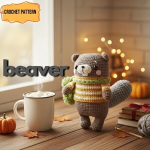 May include: A crocheted beaver doll wearing a striped sweater and a green scarf. The word "beaver" is displayed in large, gray letters. A mug of hot chocolate, a small pumpkin, and fall leaves are on a wooden surface.