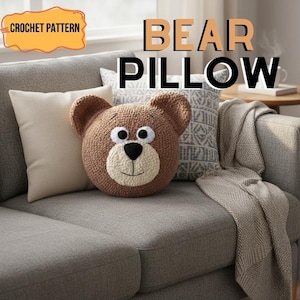 Könnte beinhalten: Ein braunes gehäkeltes Bärenkissen mit cremefarbener Schnauze und schwarzen Augen liegt auf einem grauen Sofa. Das Kissen hat eine runde Form und ist von anderen Kissen und einer Decke umgeben. Die Wörter "CROCHET PATTERN" und "BEAR PILLOW" sind sichtbar.