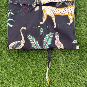 以下が含まれることがあります： フラミンゴ、ヒョウ、オウムが描かれた、カラフルなジャングルプリントの黒い布製バッグ。バッグには白いトリムと紐留めが付いています。背景は緑色です。