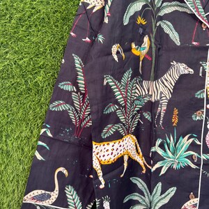 以下が含まれることがあります： 鮮やかなジャングルプリントが施された濃いネイビーのパジャマトップ。シマウマ、ヒョウ、フラミンゴ、オウムが緑豊かな葉の中に描かれています。白いパイピングと長袖が付いています。