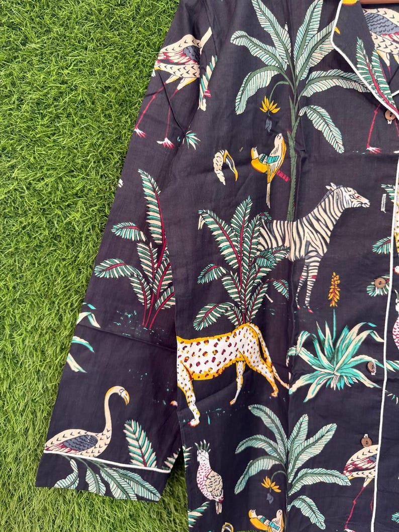 以下が含まれることがあります： 鮮やかなジャングルプリントが施された濃いネイビーのパジャマトップ。シマウマ、ヒョウ、フラミンゴ、オウムが緑豊かな葉の中に描かれています。白いパイピングと長袖が付いています。