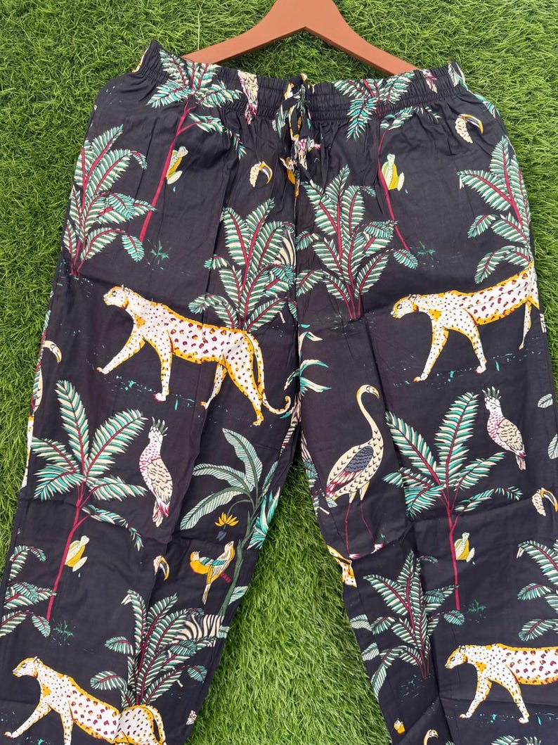 以下が含まれることがあります： ジャングルをテーマにしたプリントの黒いパジャマパンツ。ヒョウ、オウム、ヤシの木、その他の熱帯要素が含まれています。パンツはドローストリングウエストで、軽量の生地で作られています。