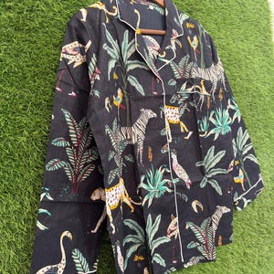 以下が含まれることがあります： 黒色のパジャマトップ。ジャングルをテーマにしたプリントで、シマウマ、ヒョウ、鳥、緑豊かな葉が描かれています。長袖のトップスは、襟、袖口、ボタンの前立てに白いパイピングが施されています。生地は濃い色です。