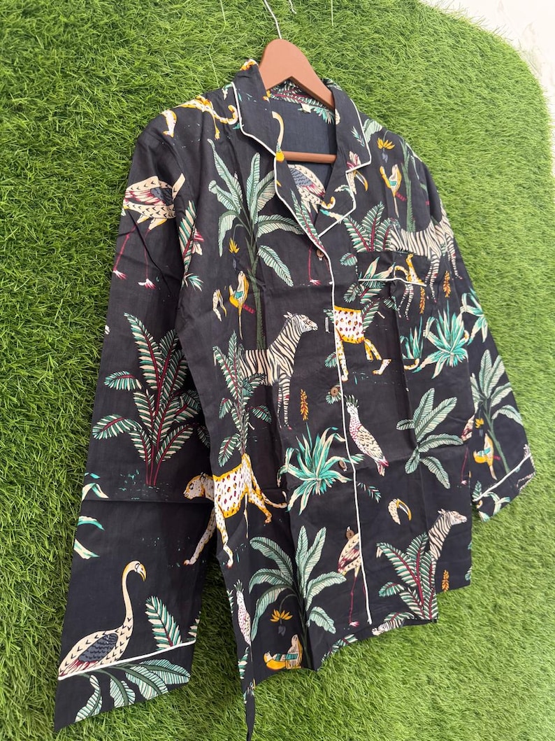 以下が含まれることがあります： 黒色のパジャマトップ。ジャングルをテーマにしたプリントで、シマウマ、ヒョウ、鳥、緑豊かな葉が描かれています。長袖のトップスは、襟、袖口、ボタンの前立てに白いパイピングが施されています。生地は濃い色です。