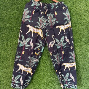 以下が含まれることがあります： ジャングルをテーマにしたプリントの黒いパジャマパンツ。デザインは、ヒョウ、オウム、ヤシの木、その他の熱帯要素を白、黄、緑、赤の色合いで表現しています。パンツは、緑色の背景に木製のハンガーに吊るされています。