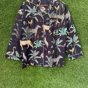 以下が含まれることがあります： 白のトリムと鮮やかなジャングルプリントが施された黒のパジャマトップ。長袖のトップスには、シマウマ、ヒョウ、鳥、そして緑豊かな葉が描かれています。ボタンダウンシャツは、緑色の背景に木製のハンガーに掛けられています。