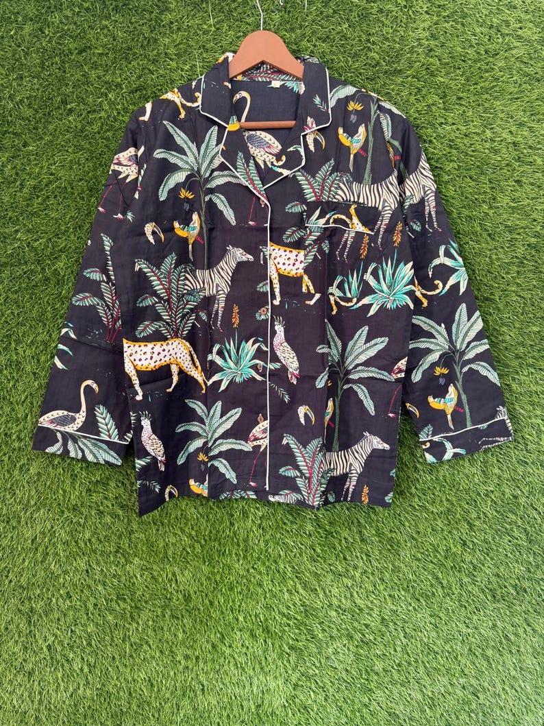 以下が含まれることがあります： 白のトリムと鮮やかなジャングルプリントが施された黒のパジャマトップ。長袖のトップスには、シマウマ、ヒョウ、鳥、そして緑豊かな葉が描かれています。ボタンダウンシャツは、緑色の背景に木製のハンガーに掛けられています。