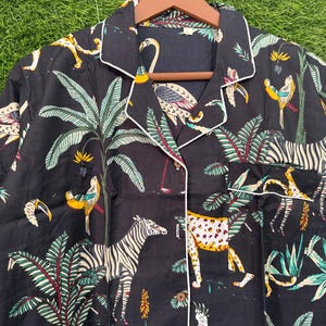 以下が含まれることがあります： 白のトリムとジャングルアニマルプリントが施された黒のパジャマトップ。プリントには、シマウマ、ヒョウ、鳥、ヤシの木が黄色、緑、白の色合いで描かれています。トップはボタンダウンで、襟が付いています。
