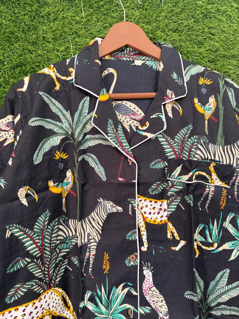 以下が含まれることがあります： 白のトリムとジャングルアニマルプリントが施された黒のパジャマトップ。プリントには、シマウマ、ヒョウ、鳥、ヤシの木が黄色、緑、白の色合いで描かれています。トップはボタンダウンで、襟が付いています。