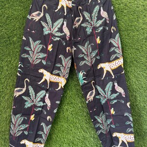 以下が含まれることがあります： ヒョウ、フラミンゴ、熱帯植物の繰り返し模様が施された黒いパジャマパンツ。パンツの裾には白い縁取りがあり、木製のハンガーに吊るされています。生地はコットンに見えます。